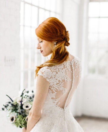 Red-haired bride with a braided low side updo, lace open-back gown and white bouquet