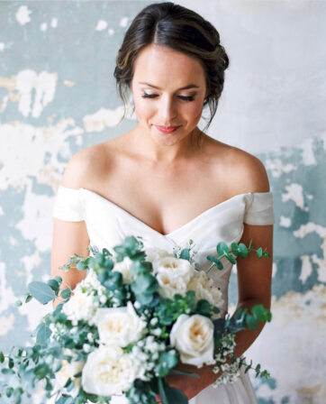 Brunette bride with a polished side-swept updo and off-shoulder gown holding white roses