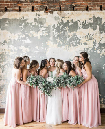 Bride and bridesmaids in blush pink dresses holding greenery bouquets by a brick wall