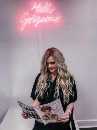 Woman with long wavy blonde hair reading a magazine under a pink Hello Gorgeous neon sign