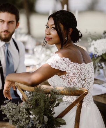 Bride with a sleek dark updo in an off-shoulder lace gown, seated at her reception