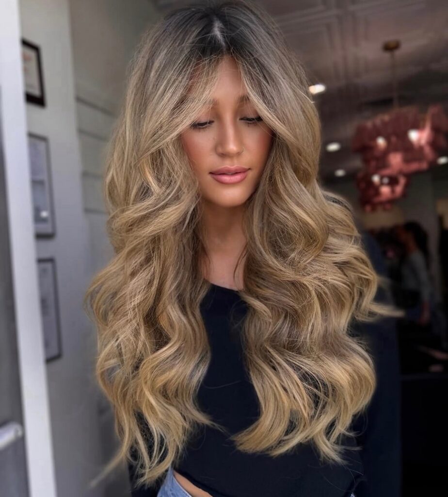 Long light honey blonde hair with a soft shadow root, curtain bangs and big, voluminous waves